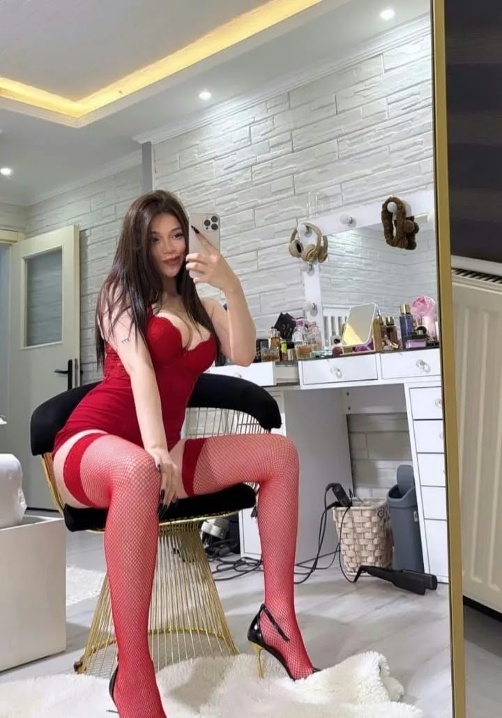 Person in red lingerie and fishnet stockings sits on a gold chair, taking a selfie in a makeup vanity with cosmetics on the desk.